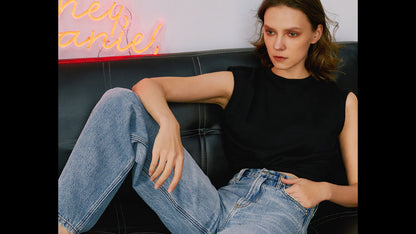 Woman sitting on a black leather couch wearing a black top and blue jeans, with a neon sign in the background.