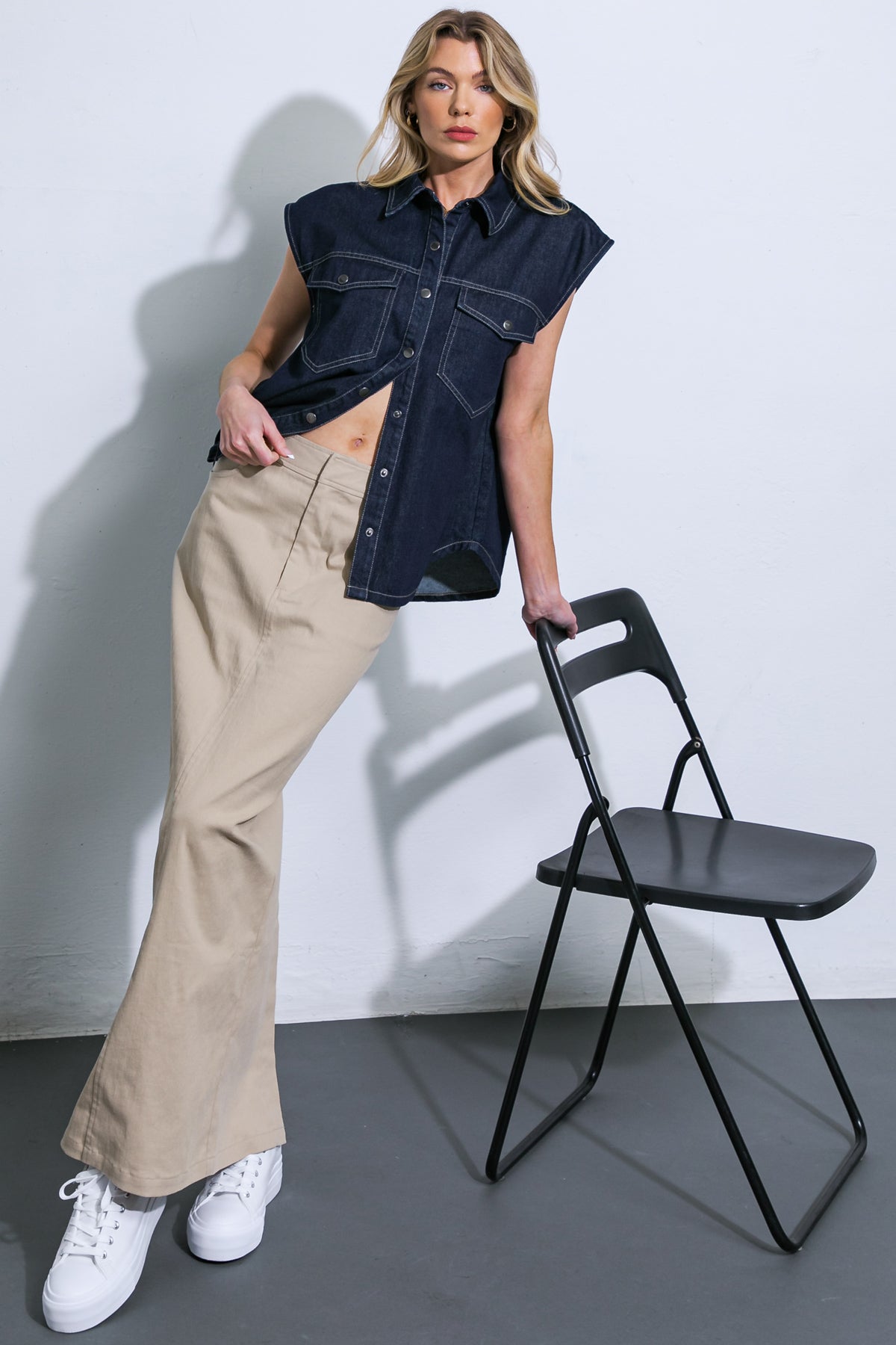 Woman wearing a denim shirt and beige pants standing next to a black chair against a white wall.